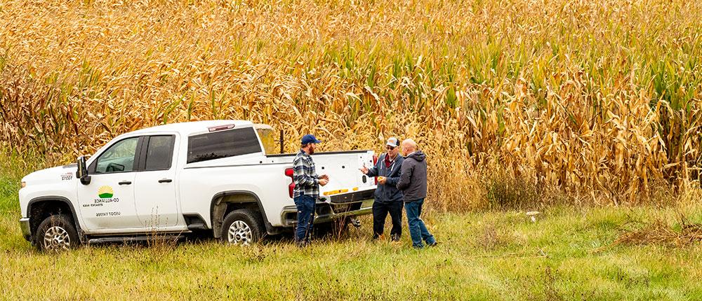 三个人在一辆卡车旁谈话，卡车侧面有联合联盟的标志, 背景是一片玉米田